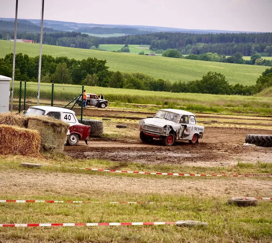 Weißer Trabant mit der Startnummer 61 auf der Schlammstrecke neben Strohballen, im Hintergrund grüne Hügel