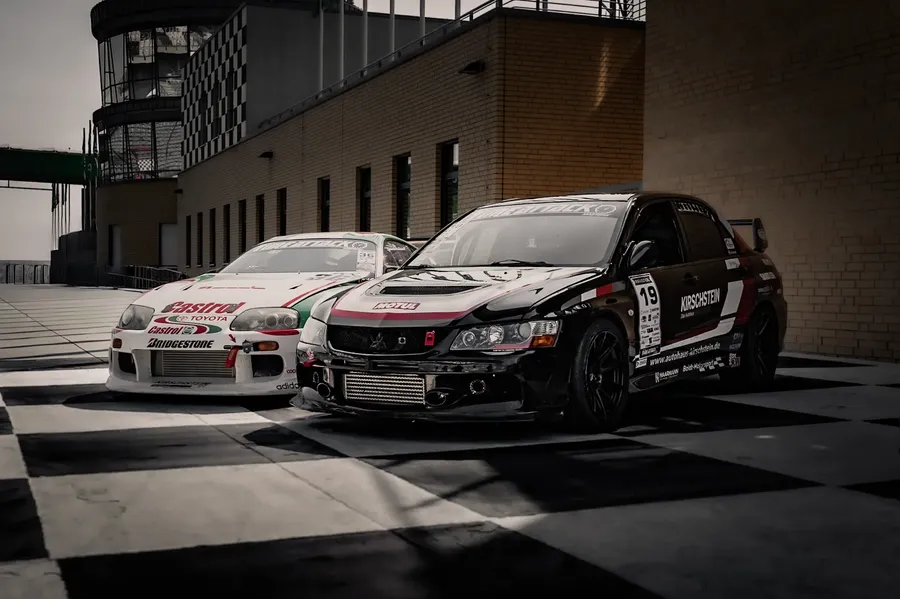 Toyota Supra MK4 in Castrol-Lackierung und schwarzer Mitsubishi Evo mit der Startnummer 19 nebeneinander auf dem Boxenvorplatz